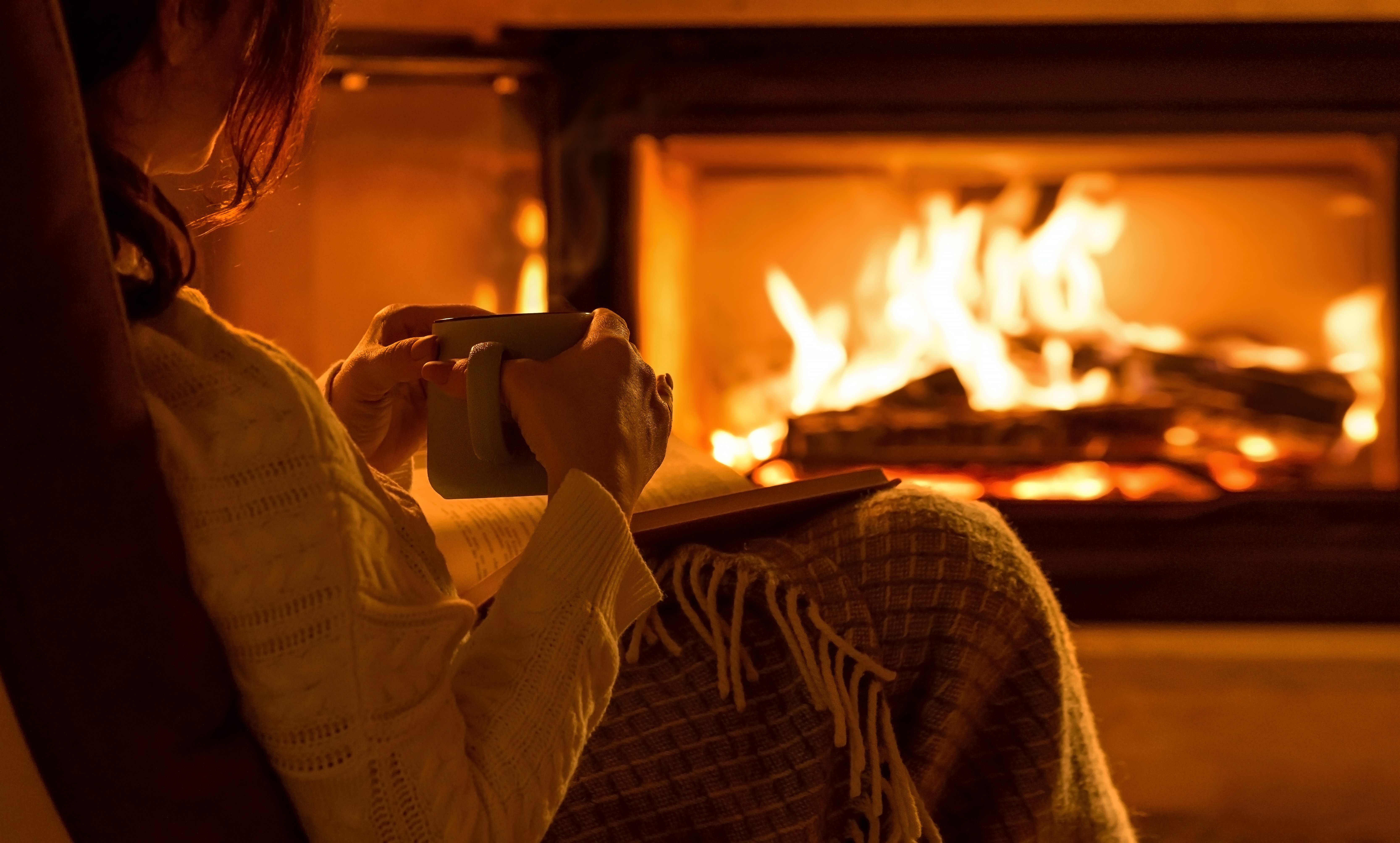 Woman wrapped in blanket holding mug by fireplace.